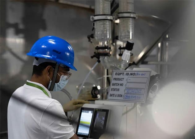 Operator in a blue hard hat and mask checking a control tablet beside a glass reactor in an API plant