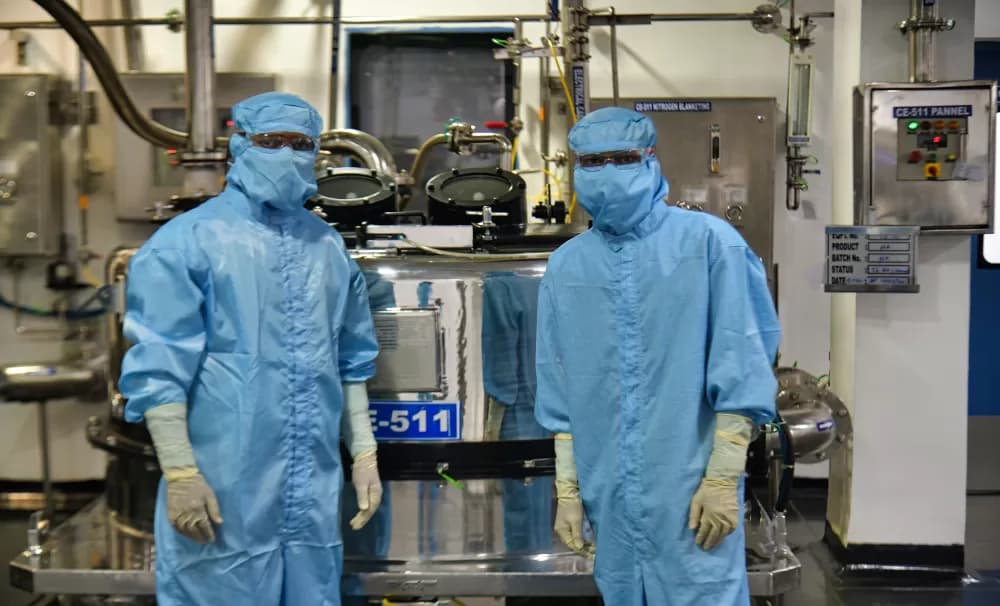 Two operators in full blue cleanroom suits standing either side of a stainless steel reactor in a pharmaceutical manufacturing plant
