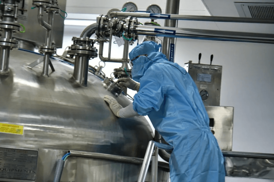 Operator in a blue cleanroom suit, hood and goggles checking a stainless steel reactor vessel at a boutique CDMO facility