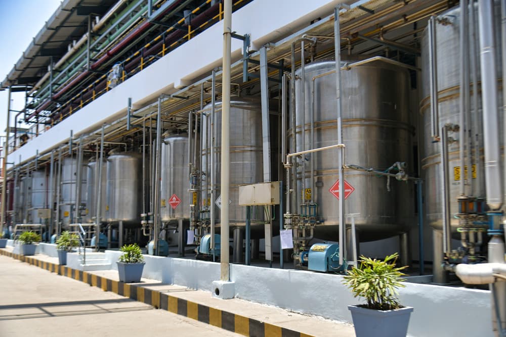 Row of large stainless steel storage tanks with pipework and hazard labels outside an API manufacturing facility block