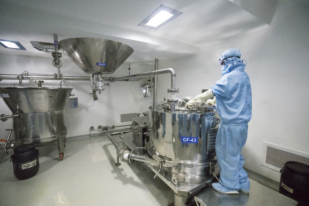 Operator in a full blue cleanroom suit working at a stainless steel centrifuge and hopper in an API processing area, illustrating micronization