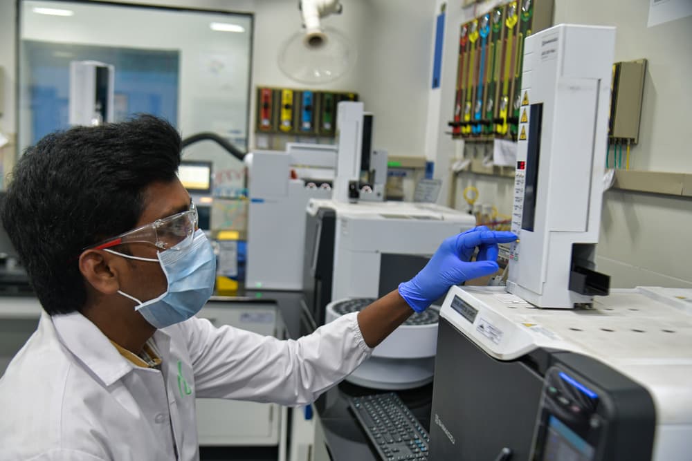Lab analyst in safety glasses, mask and blue gloves operating a gas chromatography autosampler, illustrating testing for genotoxic impurities