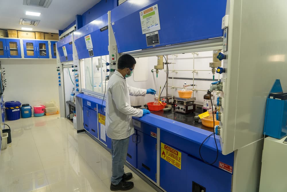 Chemist in lab coat, mask and gloves working at a fume hood in a process development lab, illustrating process engineering