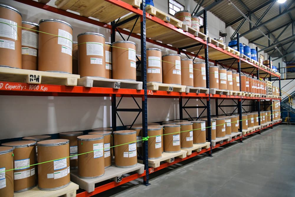 Fibre drums labelled "RM" stacked on pallet racks in a raw material warehouse, illustrating storage of regulatory starting materials