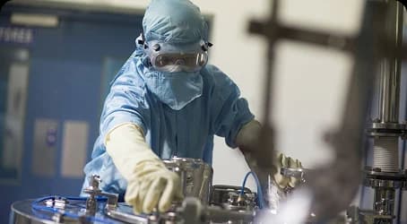 Operator in a blue cleanroom suit, hood and goggles working on a stainless steel reactor in a cGMP peptide plant