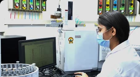 Analyst in a lab coat and face mask reviewing data on a computer next to analytical instruments