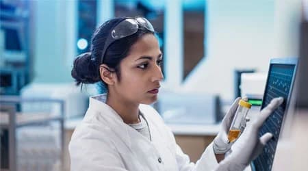 Scientist in a lab coat and safety glasses holding a sample vial while reviewing data on a tablet